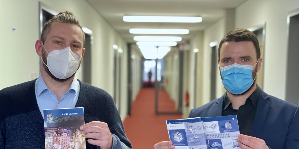 Herr Heise und Her Koppe stehen im Gang eines Verwaltungsgebäudes und präsentieren beide die Jubiläumsbroschüre.