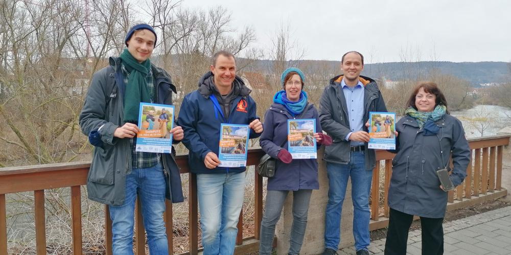 5 Menschen halten Plakate in die Kamera, hinter ihnen ein Fluss