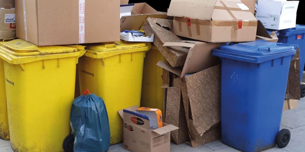 Ein Haufen Kartons steht neben, auf und zwischen gelben und blauen Abfalltonnen.