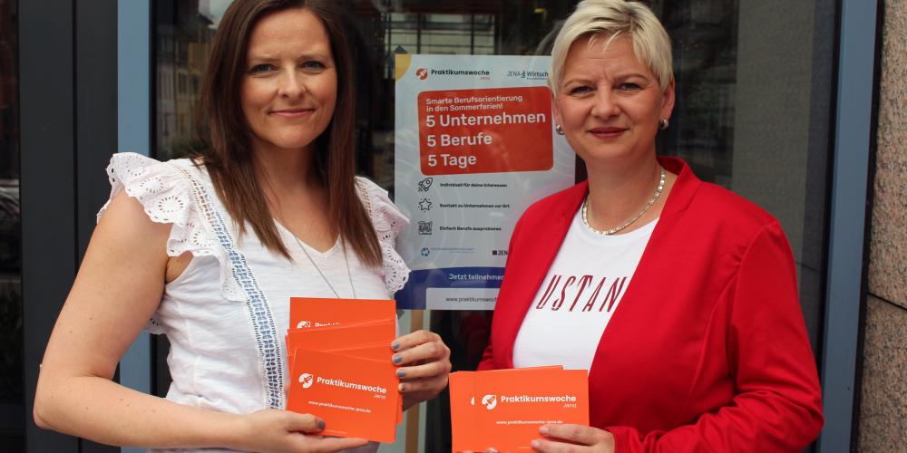 Zwei Frau stehen vor einer Glastür und schauen in die Kamera. Sie halten Flyer in der Hand. 