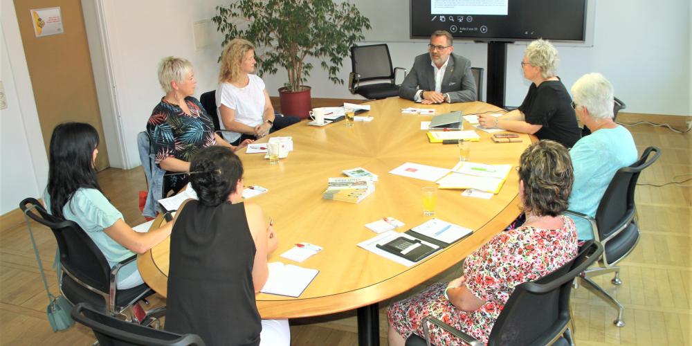 Um einen ovalen Tisch sitzen eine Mann und sieben Frauen.