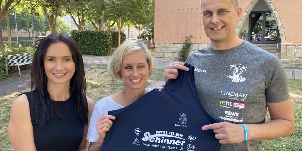 Drei Menschen lächeln in die Kamera und halten ein Oberteil mit Logos von Sponsoren hoch