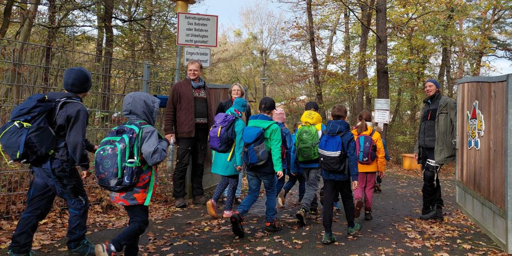 Grundschulkinder gehen durch ein geöffnetes Tor. 3 Erwachsene empfangen Sie.