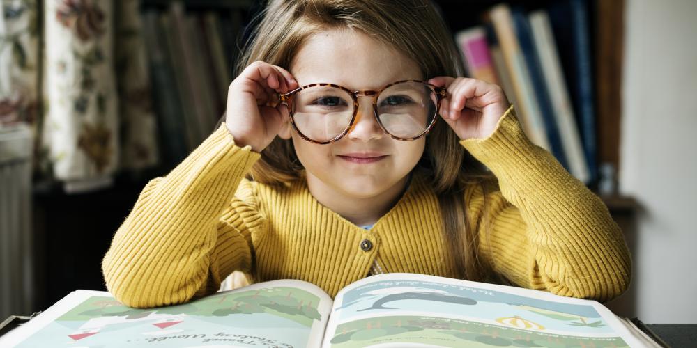 Ein Mädchen mit Brille sitzt an einem Tisch, vor ihr liegt ein großes, aufgeschlagenes Buch.