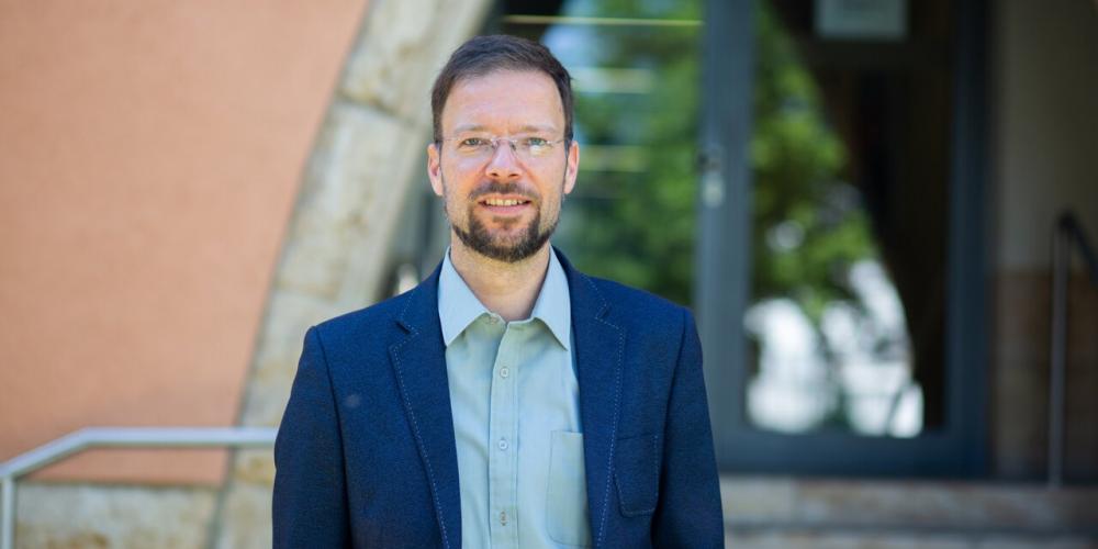 Ein Mann mit Bart und Brille steht vor dem Eingang eines Gebäudes