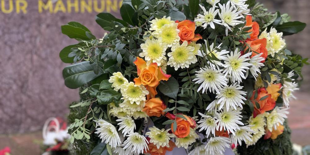 Ein Kranz mit Blumen vor einem Denkmal im Hintergrund