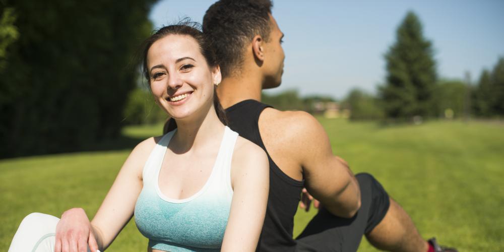 Eine junge Frau und ein junger Mann sitzen Rücken an Rück auf einer Wiese im Park. Sie tragenSportoutfits. Die Frau lächelt in die Kamera.