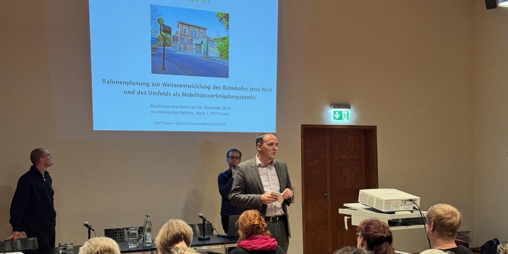 Bürgermeister Christian Gerlitz stehtim Plenarsaal im Historischen Rathaus und spreicht vor sitzenden Personen. HInter ihm ist eine Präsentation via Beamer an die Wand geworfen zum Thema Rahmenplanung zur Weiterentwicklung des Bahnhofes Jena West