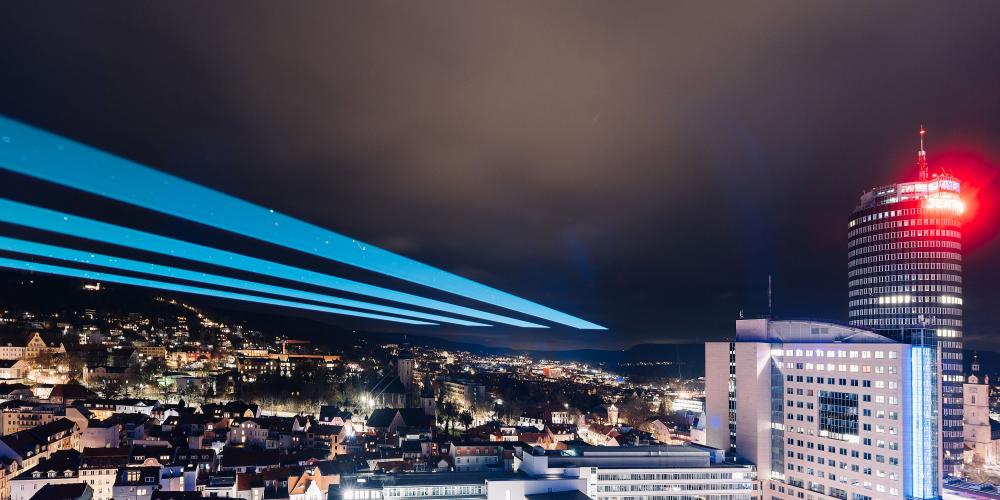 Das Foto zeigt Jena im Dunkeln mit erleuchteten Gebäuden und einem großen Laserstrahl am Himmel.
