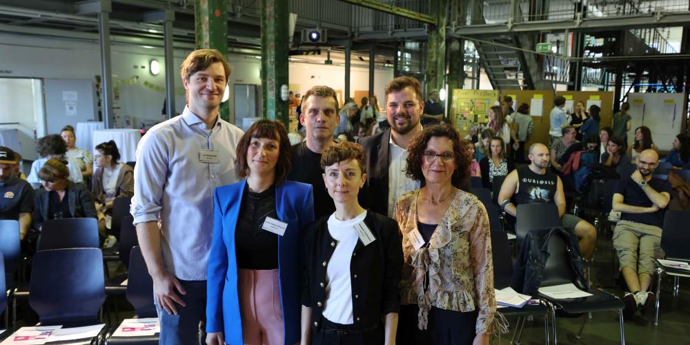 Mehrere Personen schauen in die Kamera, im Hintergrund mehrere Menschen.