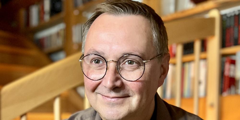 Ein Mann um die 50 sitzt vor einer Holztreppe, trägt eine Brille und ein hellbraunes Hemd.