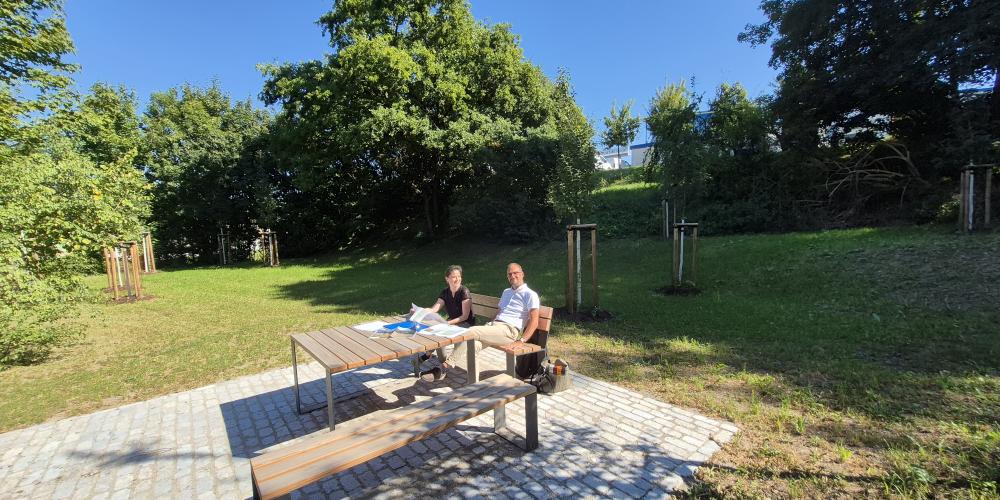 Naturnahe Lichtung mit Rastplatz: Eine Frau und ein Mann sitzen in der Sonne auf einer Bank.