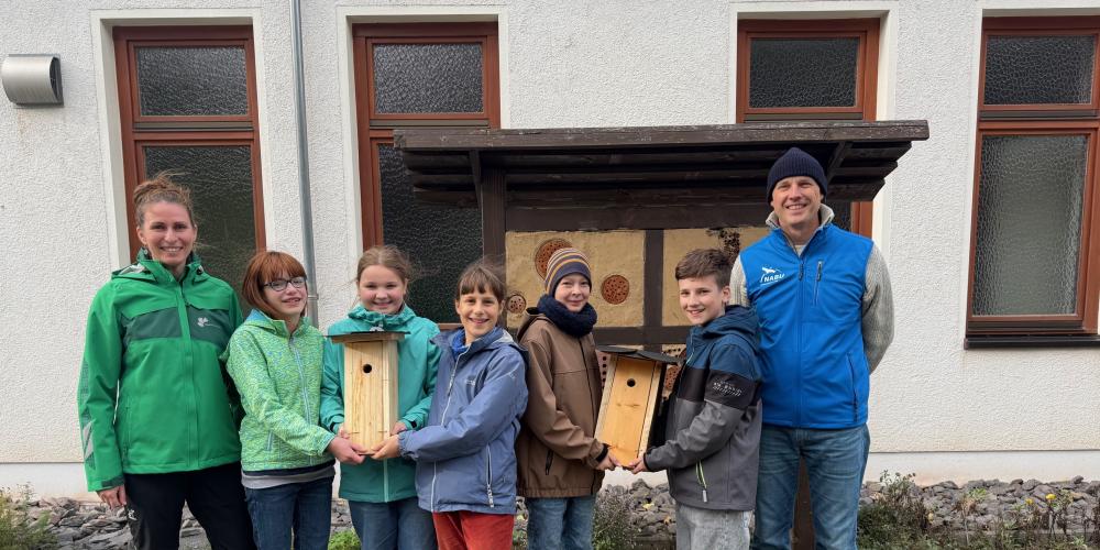 Fünf Jugendliche (drei Mädchen und zwei Jungen) halten zwei Nistkästen aus Holz. Links neben ihnen steht eine Frau und auf der rechte Seite ein Mann.