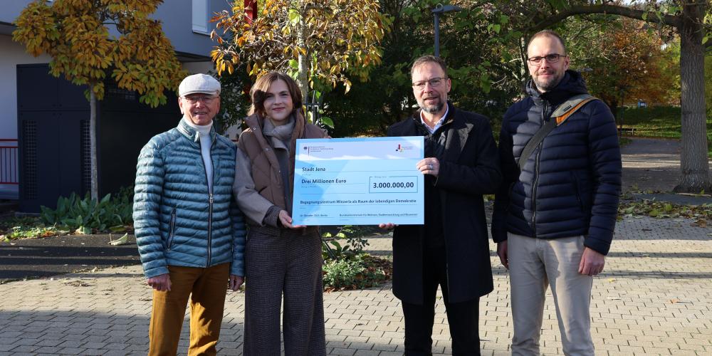 v.l.: Friedrich Wilhelm Gebhardt, ehemaliger Ortsteilbürgermeister von Jena-Winzerla, Ostbeauftragte Elisabeth Kaiser, Oberbürgermeister Thomas Nitzsche und Lars Liebe, Fachdienstleiter Stadtentwicklung stehen nebeneinander und halten einen großen Scheck.