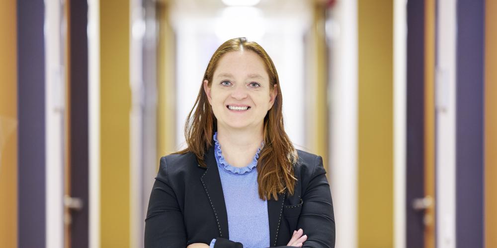 Frau steht mit verschränkten Armen in einem Korridor und lächelt in die Kamera