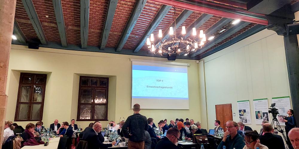 Stadtratsmitglieder sitzen an Tischen im historischen Rathaus, ein Redner steht am Mikrofon