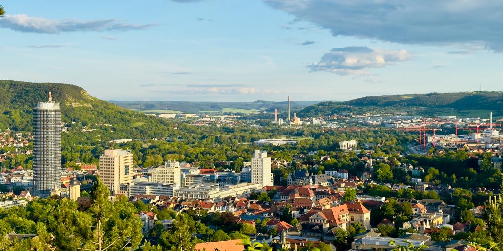 Das Foto zeigt eine weite Panoramaaufnahme der Stadt Jena in Thüringen, aufgenommen vom Landgrafenblick aus. Im Vordergrund sind grüne Baumkronen zu sehen, dahinter breitet sich die Stadt mit ihren Wohnhäusern und Bürogebäuden aus. Links ragt der runde Jentower deutlich über die übrigen Gebäude hinaus. Im Hintergrund ziehen sich die bewaldeten Hügel des Saaletals entlang, und der Himmel ist leicht bewölkt mit Sonnenlicht, das die Landschaft in warmes, freundliches Licht taucht.