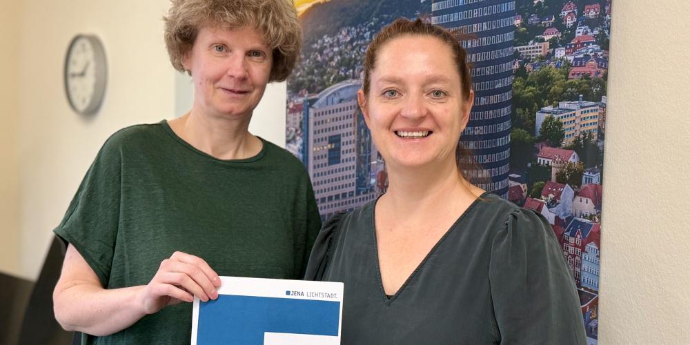 Zwei Frauen schauen freundlich in die Kamera und halten eine Broschüre