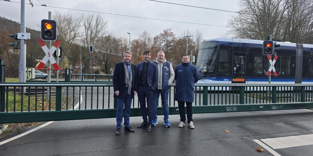 Vier Männer stehen auf der Straße vor einer geschlossenen Schranke. Im Hintergrund fährt eine Straßenbahn vorbei. 