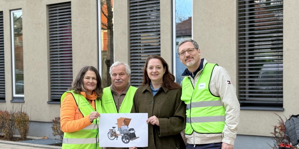 Vier Personen stehen nebeneinander vor einem modernen Gebäude mit großen Fenstern. Drei von ihnen tragen leuchtend gelbe Warnwesten, eine Person in der Mitte trägt eine dunkelgrüne Jacke. Die beiden Personen links halten gemeinsam ein Foto einer Rikscha, die für das Projekt »Radeln ohne Alter« vorgesehen ist. Alle lächeln in die Kamera.