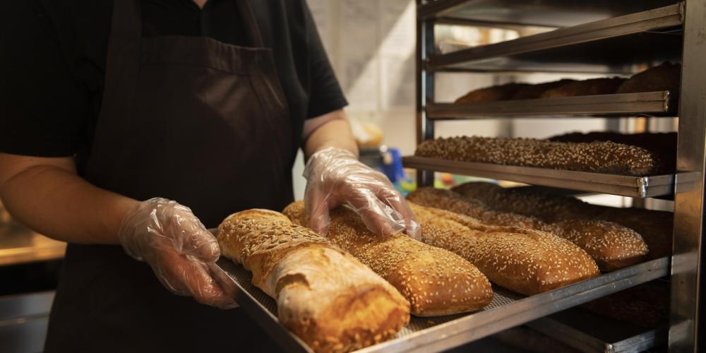 Eine Person in dunkler Arbeitskleidung und transparenten Einweghandschuhen hält ein Blech mit frisch gebackenen, langen Broten, teilweise mit Sesam bestreut. Im Hintergrund steht ein Metallregal mit weiteren Broten in einer Backstube.