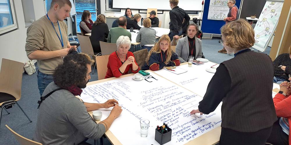 In einem hellen Seminarraum sitzen mehrere Gruppen von Menschen an Tischen und arbeiten gemeinsam an großen Papierbögen. Im Vordergrund beugen sich Teilnehmende über ein großflächiges Blatt, auf dem viele handgeschriebene Stichpunkte stehen. Eine Frau zeigt auf einzelne Notizen, während andere aufmerksam zuhören. Im Hintergrund sind weitere Kleingruppen im Gespräch, Moderationsmaterialien und beschriebene Flipcharts sichtbar. Die Stimmung wirkt konzentriert und kooperativ.
