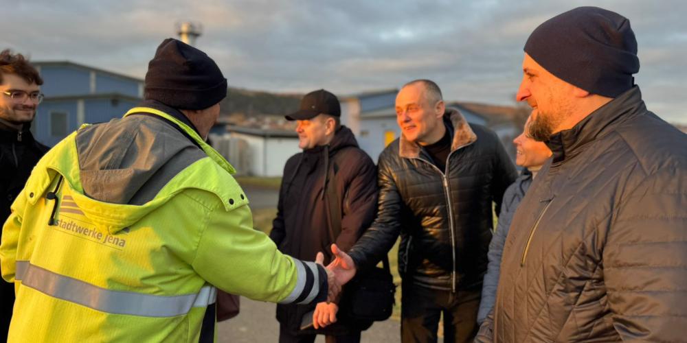 Eine Person in einer gelben Jacke der Stadtwerke Jena gibt mehreren Besuchern im Freien die Hand. Die Gruppe steht lächelnd beisammen, im Hintergrund sind Gebäude und ein bewölkter Himmel zu sehen, das Licht der tiefstehenden Sonne fällt warm auf die Szene.
