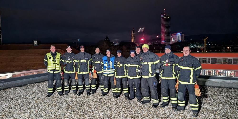 Mehrere Feuerwehrmänner schauen in die Kamera, im Hintergrund Feuerwerk.