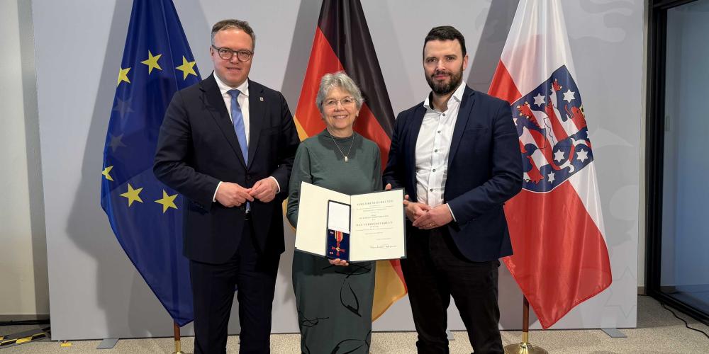 Drei Menschen stehen vor Fahnen und schauen in die Kamera.