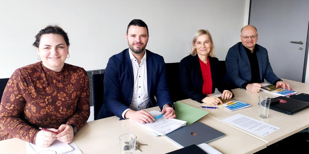 Zwei Frauen und zwei Männer sitzen an einem Tisch, vor ihnen sind Unterlagen ausgebreitet, sie lächeln in die Kamera.
