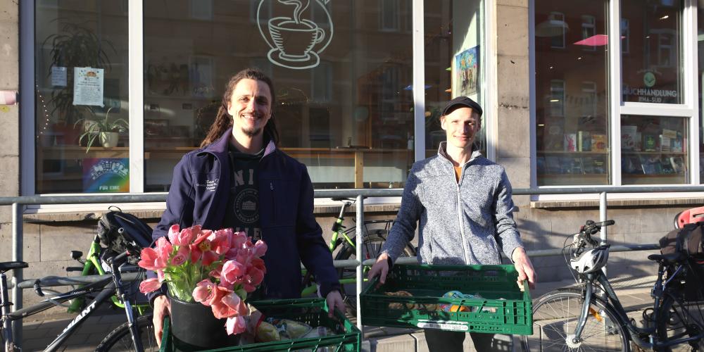 Zwei Männer stehen vor einem Geschäft und halten je eine große Klappkiste mit Lebensmittel und Blumen in der Hand