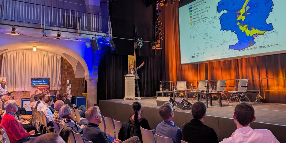 Auf einer Bühne stellt ein Mann eine Präsentation vor. Personen sitzen davor auf Stühlen in mehreren Reihen und hören zu.