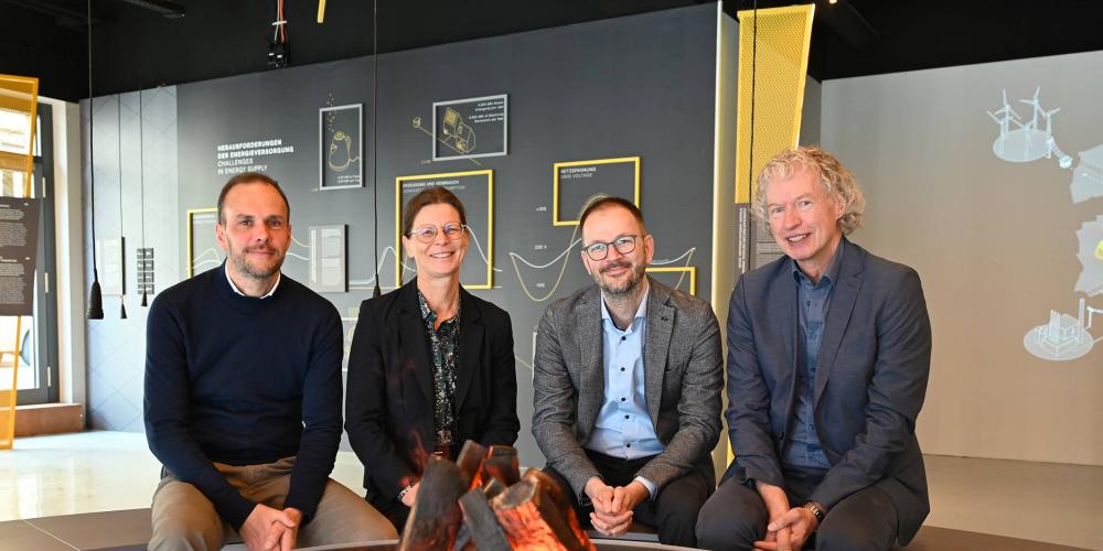 Drei Männer und eine Frau sitzen an einem künstlichen Lagerfeuer in einer Ausstellung.