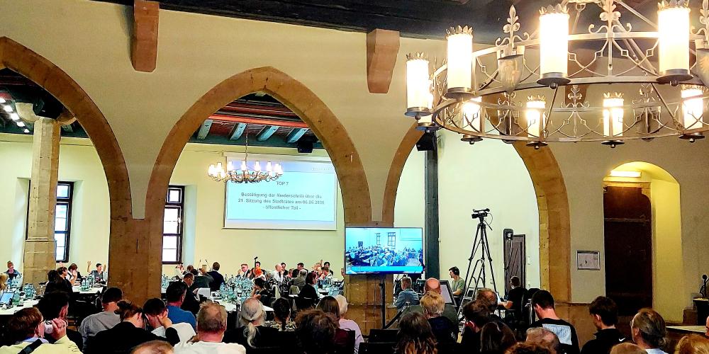 Saal des historischen Rathauses während der Stadtratssitzung mit Menschen auf Stühlen und an Tischen sitzend
