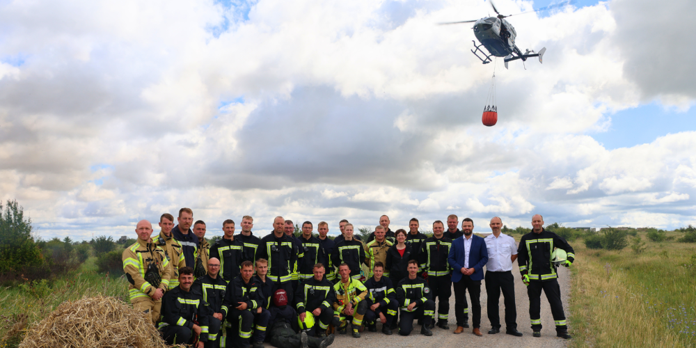 Mehrere Männer stehen auf einem weiten Wiesenfeld, ein Hubschrauber im Hintergrund.