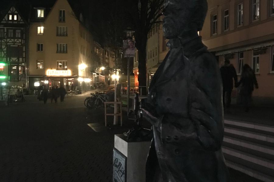 Ein Denkmal eines Mannes auf einem dunklem Platz, im Hintergrund Laternen einer Straße