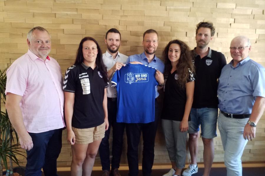 Fünd Männer und zwei Frauen halten ein Fußball-Trikot in die Kamera.