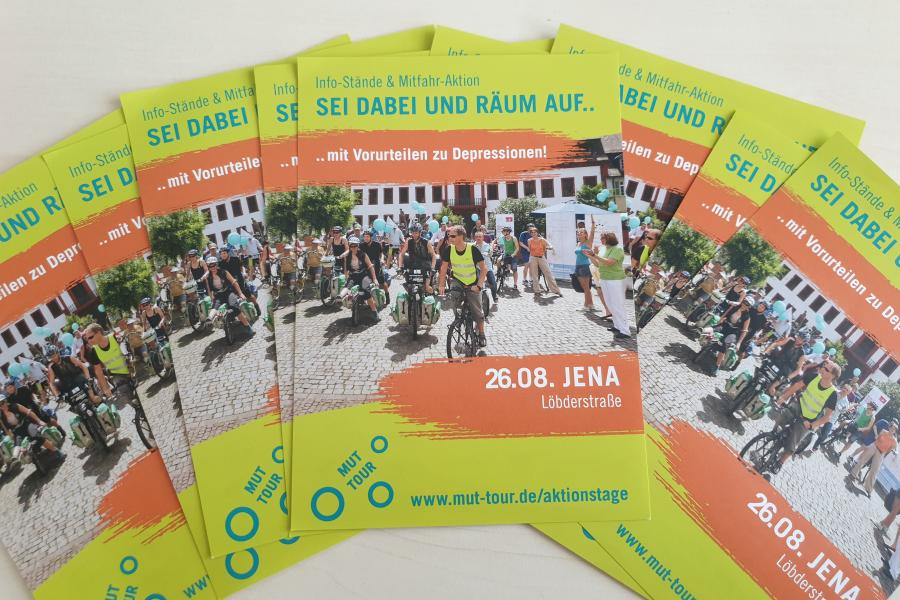 Mehrere Flyer der MUT-Tour liegen aufgefächert auf einem Tisch. Es ist ein Bild mit Fahrradfahrern zu sehen und der Termin in Jena.