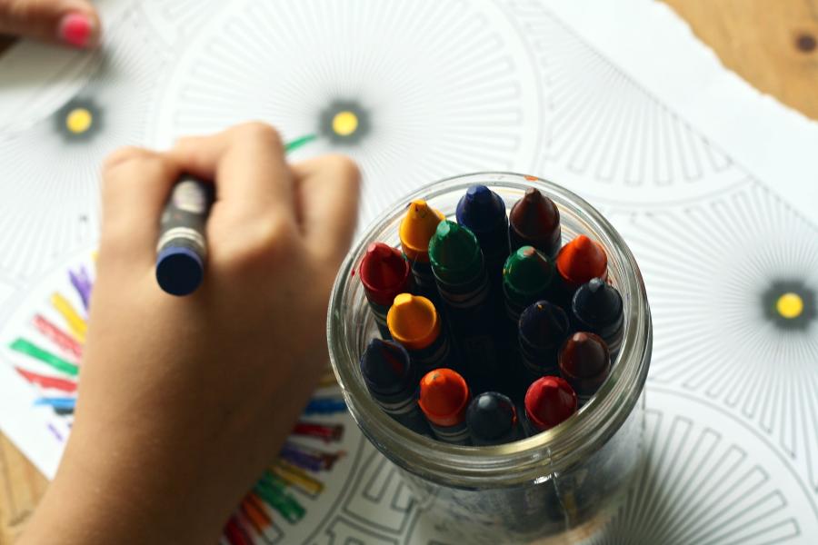 Kinderhand mit Wachsmalstift malt auf einem Stück Papier, dass auf einem Tisch liegt, daneben mehrere Wachsmalstifte in einem Glas