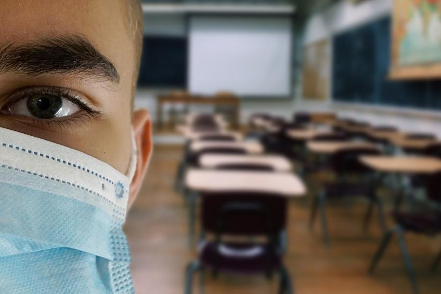 Halbes Gesicht einen Schülers mit Maske dahinter ein leeres Klassenzimmer