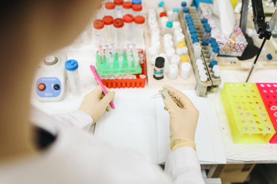 Person mit Handschuhen schreibt in einem Labor mit Testequipment etwas auf.