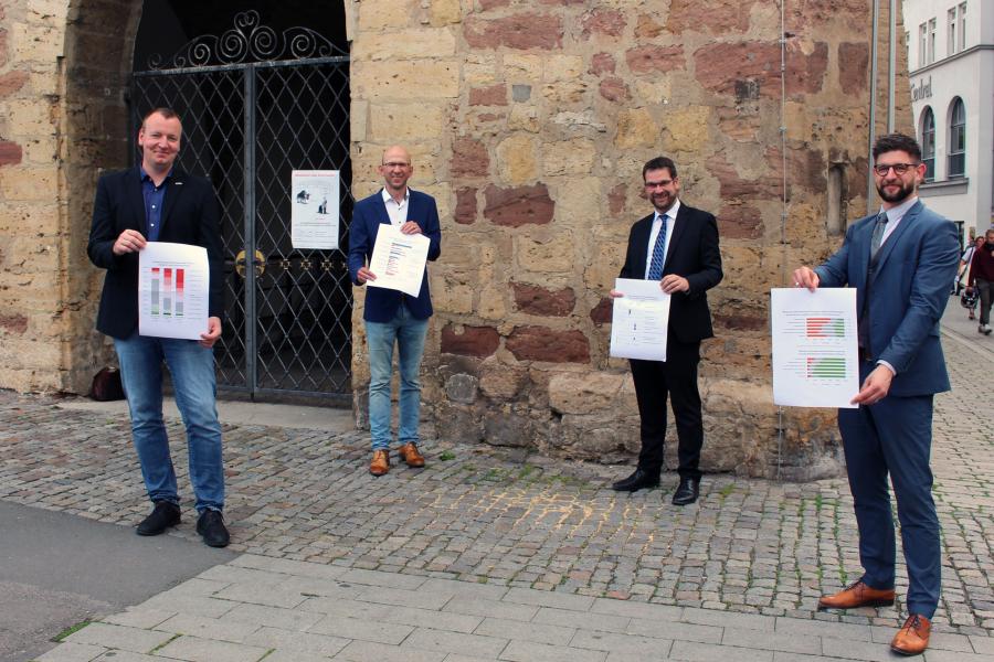 Vier Männer halten Papier in die Kamera und halten Abstand voneinander, im Hintergrund das Rathaus