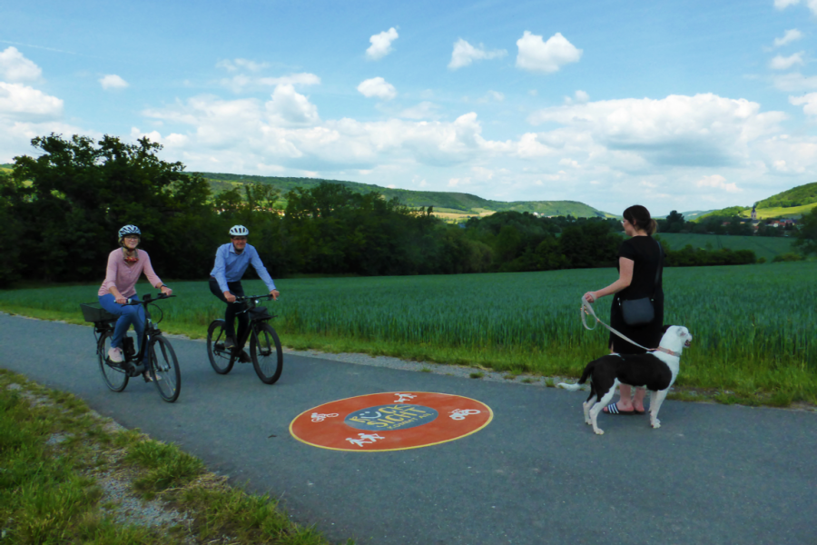Drei Menschen auf einem Fahrradweg, zwei mit Fahrrad, eine Dame mit Hund