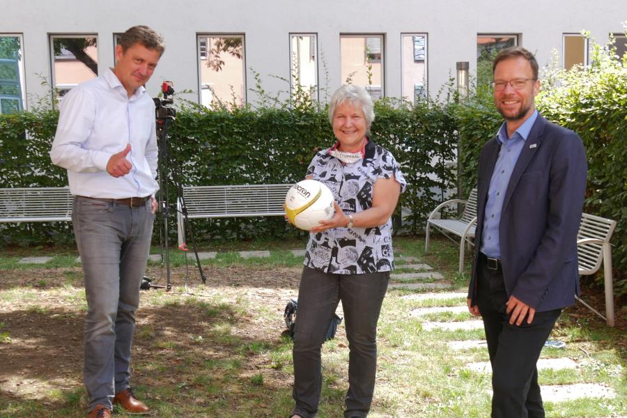 Drei Mensch schauen in die Kamera, die Frau in der Mitte hält einen Fußball.