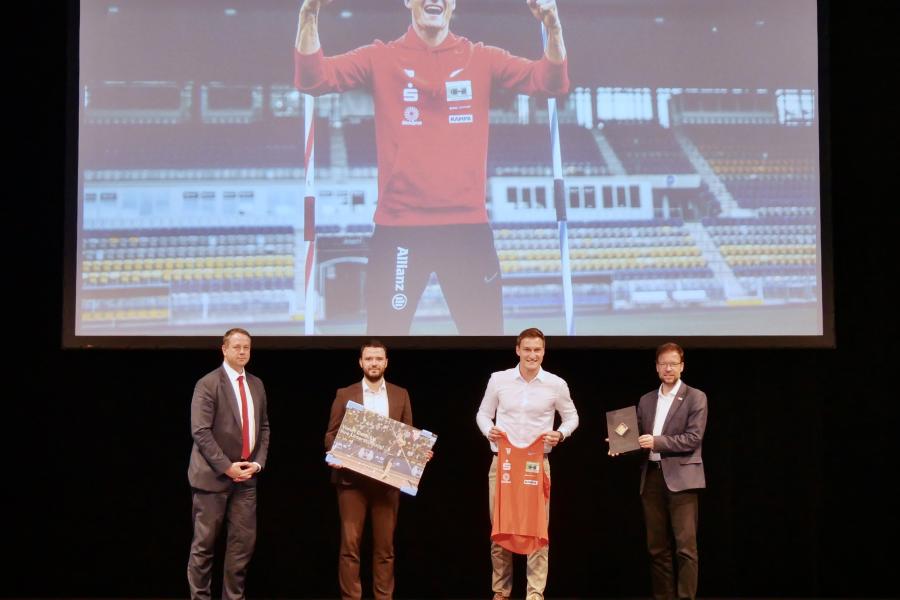 Vier Männer auf ein Bühne, im Hintergrund ein großes Bild eines Sportlers