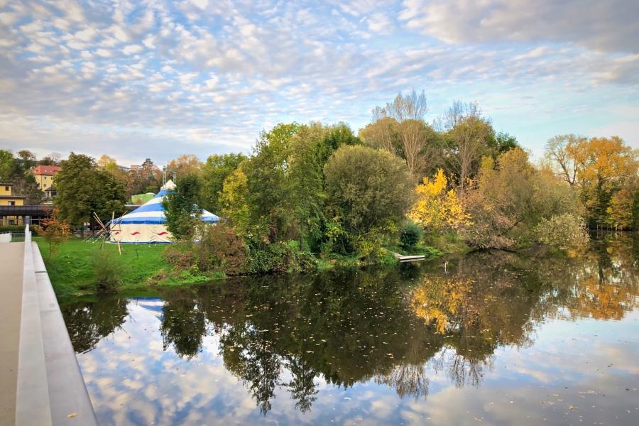 Im Vordergrund ein Fluss mit einer Brücker und im Hintergrund ein Zirkuszelt im Grünen
