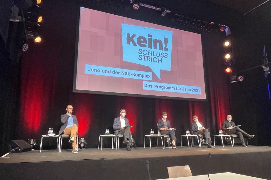 5 Personen sitzen auf Stühlen auf einer Bühne, Leinwand im Hintergrund