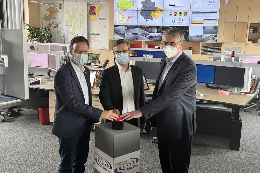 Drei Männer in einem Raum voller Computerarbeitsplätze drücken auf einen großen Knopf