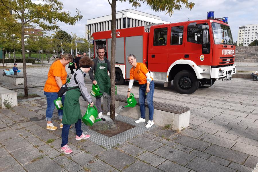 Vier Personen gießen einen Baum, im Hintergrund ein Feuerwehrwagen