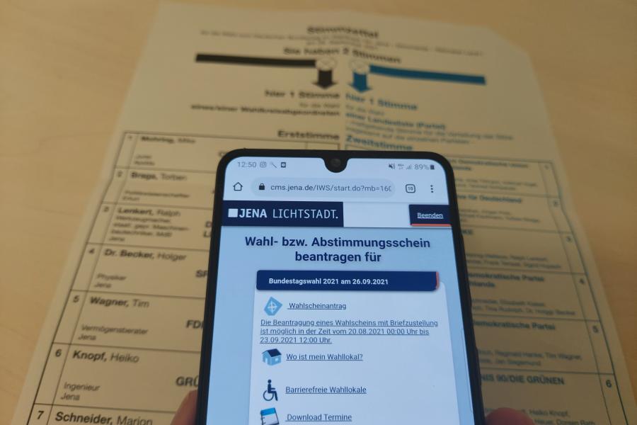Ein Wahlschein liegt auf einem Tisch, darüber ein Smartfone mit der geöffneten Seite um die Briefwahl zu beantragen.